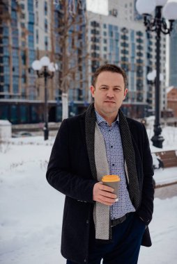Paltolu ve takım elbiseli genç bir işadamı elinde kahveyle şehrin ara sokağında duruyor.