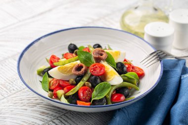 Akdeniz salatasına ançüez, yeşil fasulye, domates, yumurta, siyah zeytin, beyaz tabakta kapari ve beyaz ahşap zemin. Sağlıklı yemek, karbonhidrat diyeti yok..