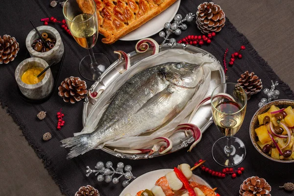 Christmas eve dinner concept. Baked dorado fish, fish pie, potato salad, seafood skewers. White wine glases. Festive decoration. Catholic tradition related to the Feast.