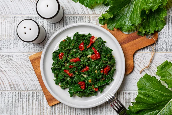 Cooked turnip greens with garlic and tomatoes. The dark leafy edible green tops of turnips. They are high in nutrients and low in calories. On white wood table surface.