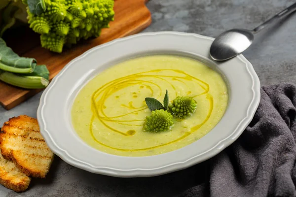 Eating creamy romanesco broccoli soup and toasted bread. Vegan dish. Fresh roman broccoli on background. Giagonal composition on dark grey surface.