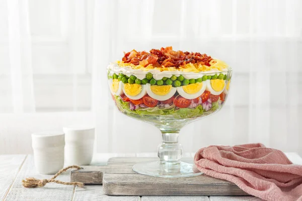 Seven-layer salad. American dish with ingredients: iceberg lettuce, tomatoes, red onions, sweet peas, hard boiled eggs, cheddar cheese, and bacon pieces topped with a mayonnaise dressing.