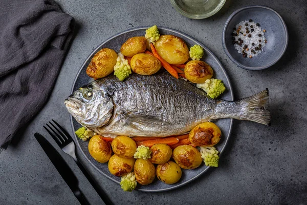 Baked whole dorado fish with baked vegetables, potatoes, carrots and romanesco cauliflower. Mediterranean dinner served on a gray plate.