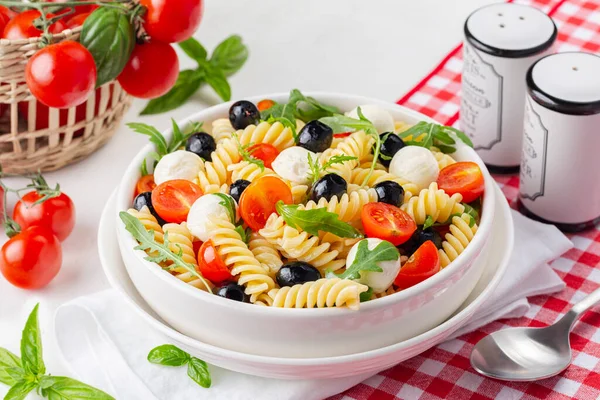 Italian cold pasta salad or Pasta fredda alla caprese. Fusilli, tomato, mozzarella, olive, arugula. Table with white bowl, fresh vegetables on background.