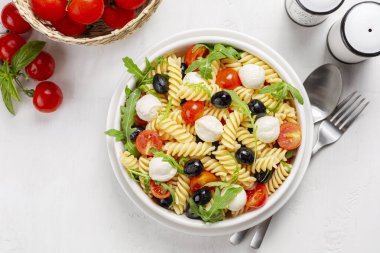 İtalyan soğuk makarna salatası. Makarna fredda alla caprese. Fusilli, domates, mozzarella, zeytin, roka. Beyaz bir masa yüzeyinin hemen üstünde..