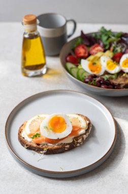 Sağlıklı kahvaltı ya da orta pişmiş yumurtalı atıştırmalık peynir kremalı ekmek, somon balığı ve gri tabakta limon dilimleri. Bir kase salata ve zeytinyağı.