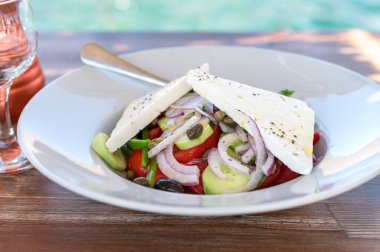 Traditional Greek Salad served in tavern, traditional greece food with Aegean sea as background. Tomatoes, cucumber, onions, olives, peppers, cappers and olive oil