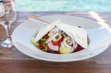 Traditional Greek Salad served in tavern, traditional greece food with Aegean sea as background. Tomatoes, cucumber, onions, olives, peppers, cappers and olive oil