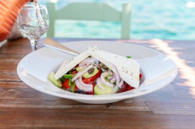 Traditional Greek Salad served in tavern, traditional greece food with Aegean sea as background. Tomatoes, cucumber, onions, olives, peppers, cappers and olive oil