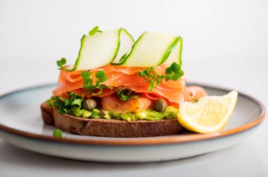 Toasted bread with mashed avocado, cucumber and smoked salmon on grey plate