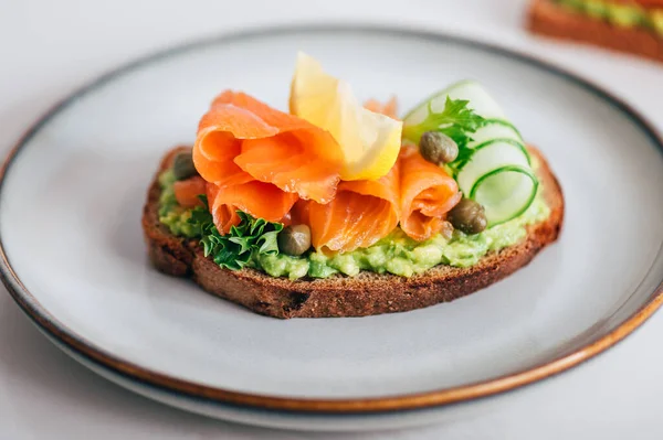 Toasted bread with mashed avocado, cucumber and smoked salmon on grey plate