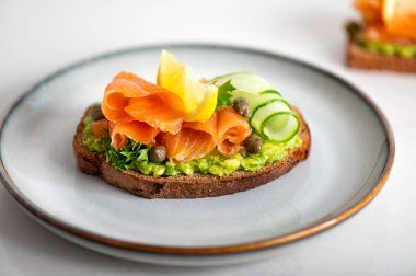 Toasted bread with mashed avocado, cucumber and smoked salmon on grey plate