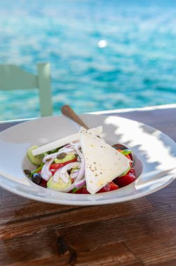 Traditional Greek Salad served in tavern, traditional greece food with Aegean sea as background. Tomatoes, cucumber, onions, olives, peppers, cappers and olive oil