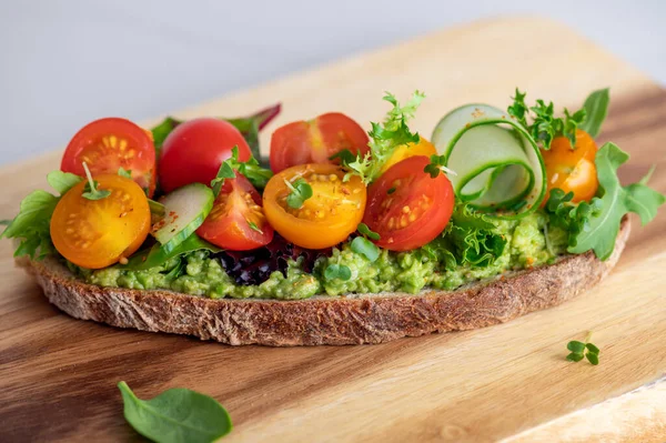 Tasty open sandwich from toasted sourdough bread with mashed avocado, slices of fresh cucumber and cherry tomatoes