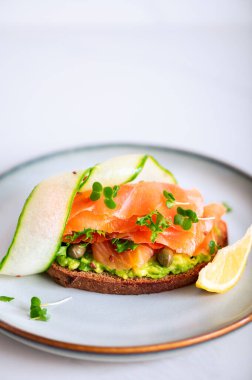 Toasted bread with mashed avocado, cucumber and smoked salmon on grey plate