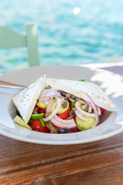 Traditional Greek Salad served in tavern, traditional greece food with Aegean sea as background. Tomatoes, cucumber, onions, olives, peppers, cappers and olive oil