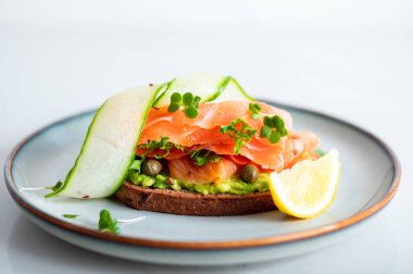 Toasted bread with mashed avocado, cucumber and smoked salmon on grey plate