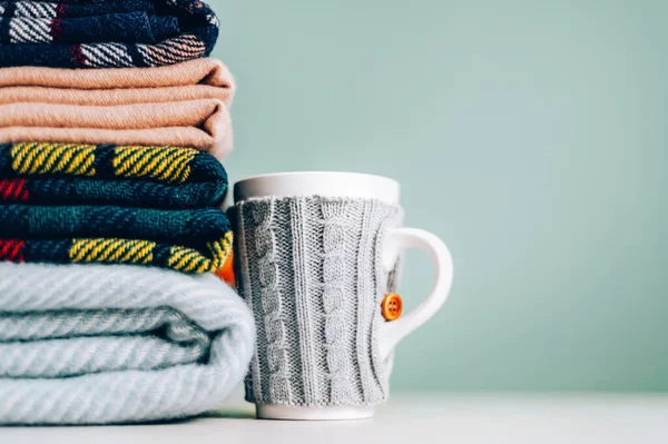 Stack of folded woolen checked blankets and mug in wollen cover, autumn and cold winter concept