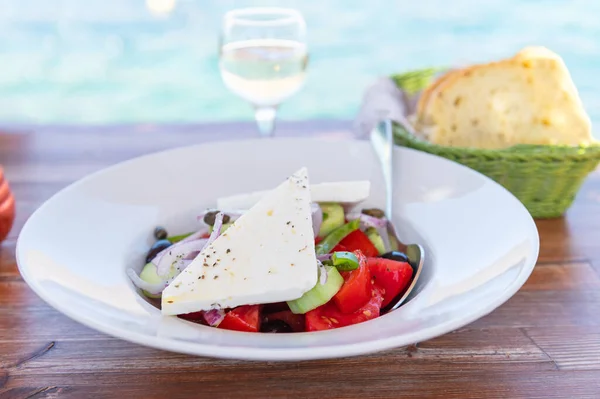 Traditional Greek Salad served in tavern, traditional greece food with Aegean sea as background. Tomatoes, cucumber, onions, olives, peppers, cappers and olive oil