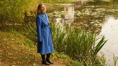 Göletin yanındaki parkta, yere düşmüş sonbahar sarısı yaprakları olan bir kadın. Mavi ceketli ve siyah çizmeli güzel bir sarışının portresi.