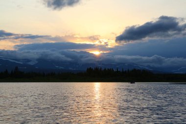 Gölün üzerinde gün batımı ve gri bulutların içinde güneşli dağlar. Suya yansıyan bir yaz günü Ural Rusya 'nın sonunun resmi.