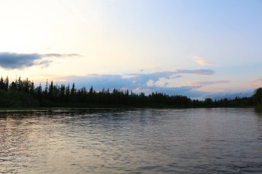 Gün batımında nehir manzarası sahilde bir ormanın silueti ve bulutlarla aydınlatılmış mavi gökyüzü.