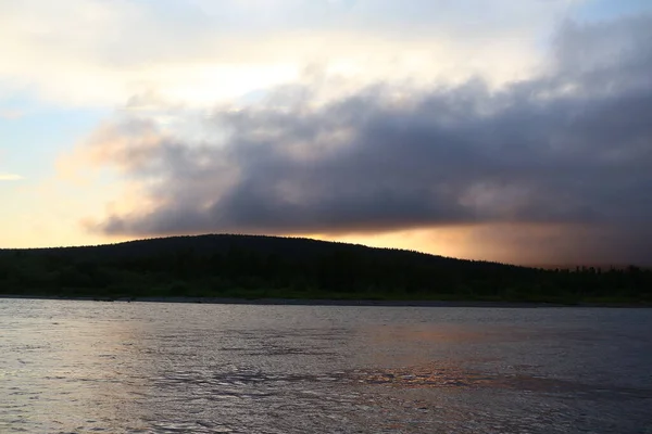 夏の夜に山の黒いシルエットの後ろに金色の光で曇り空を照らす夕日と湖の上の夕日 — ストック写真