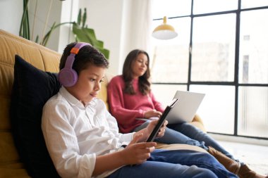 Millennial anne, genç oğluyla evden çalışıyor. Çocuk bir tabletle oynuyor. Evdeki aile hayatı ve tele çalışma kavramı.