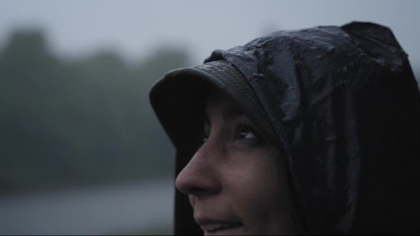 En filmisk bild av en leende kvinna som blir blöt under regnet i naturen. Hon bär regnrock. — Stockvideo