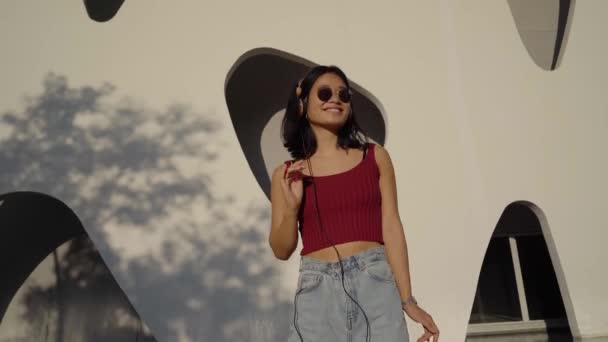 Feliz mujer asiática cool usando auriculares bailando solo en la calle. Sonriente jovencita hipster escuchando música de pie en la ciudad al aire libre, sintiéndose libre y funky. — Vídeos de Stock