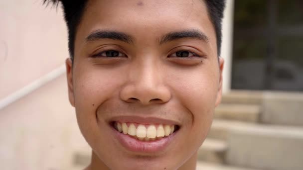Ázsiai tinédzser boldogan mosolyog. Közelkép portré vidám fiú nézi a kamera kint áll homályos háttér. — Stock videók