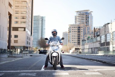 Wide shot of a young motorcyclist stopped at a traffic light in Barcelona. The man riding his scooter through the city on a large avenue lined with skyscrapers clipart