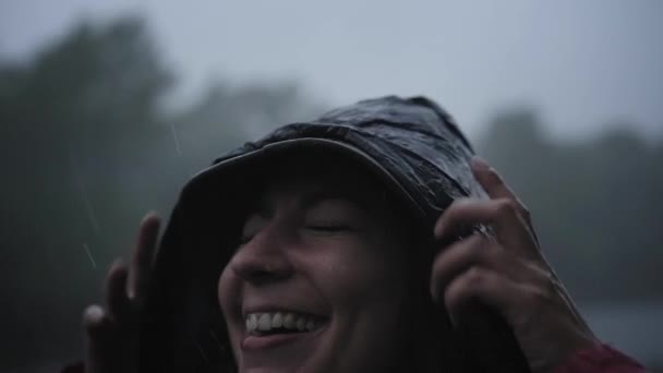 Yağmurda gülümseyen kadın yağmurluk giyerek doğanın tadını çıkarıyor. Damlalar onun yüzüne düşer ve kız mutlu olur. Doğa kavramı, özgürlük, saflık... — Stok video