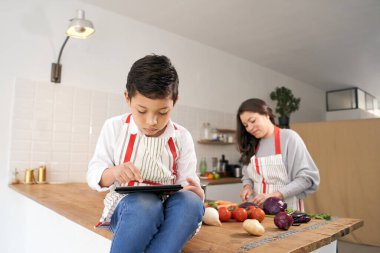 Anne sebze pişirirken önlüklü çocuk tabletle oynuyor. Sağlıklı beslenme.