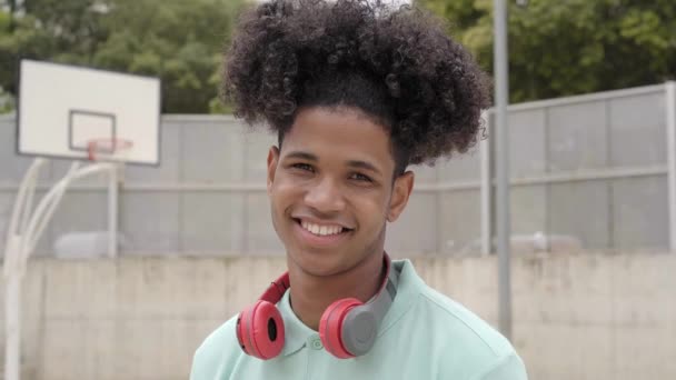 Primer plano de una sonrisa adolescente mirando una cámara feliz. Joven latino con pelo afro en una cancha de baloncesto con cascos. — Vídeo de stock