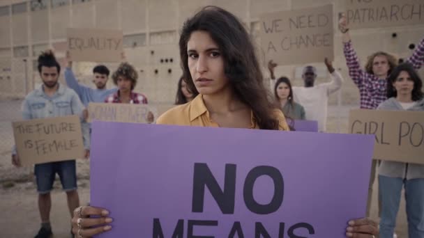 Vrouw in Demonstratie die met een serieus gezicht naar de camera kijkt. Groep mensen met bordje met woord in de buurt van feminist. — Stockvideo