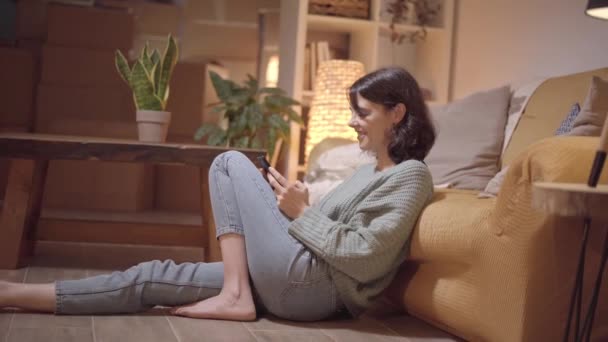 Mujer sentada en el piso del comedor un teléfono inteligente en casa. En línea desde el teléfono celular en interiores. — Vídeo de stock