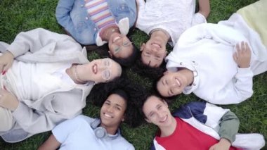 Bir grup mutlu genç çimlere uzanıp kameraya bakıyor. Gülümseyen bin yıllık arkadaşlar birlikte eğleniyorlar. Çok ırklı öğrenciler ve gençlik kültürü.