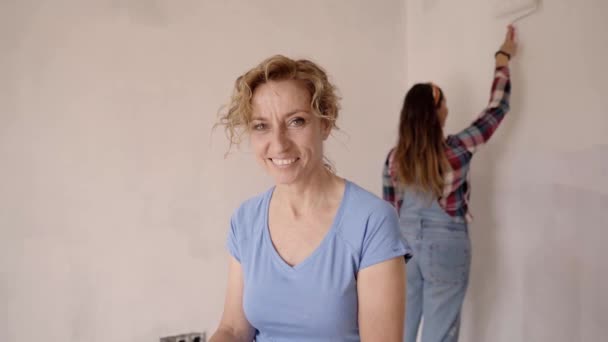 Жінка середнього віку дивиться на камеру. Друзі-жінки роблять ремонт разом, малюють білу стіну в порожній квартирі з роликами, повільний рух — стокове відео