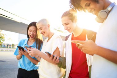 Happy millennial group of students using smartphone outdoors at city. Multi-ethnic teen people sharing in social media with her cell phones. Focus is on caucasian guy 