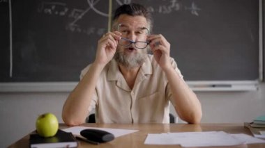 Webcam view of the teacher in online classes. Man with gray hair and beard puts on his glasses while explaining the lesson from a distance.