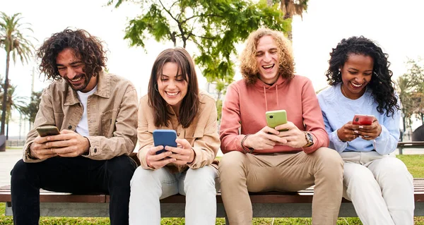 Mosolygó vegyes verseny baráti társaság ül együtt padon mobiltelefon a parkban - barátok szórakozás megosztása a közösségi média — Stock Fotó