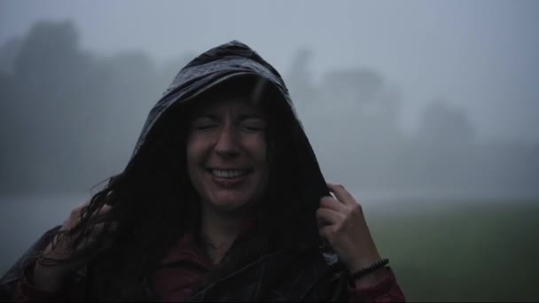若い女性は雨のシャワーの外に立っている。笑顔幸せな女の子を着てレインコート. — ストック動画