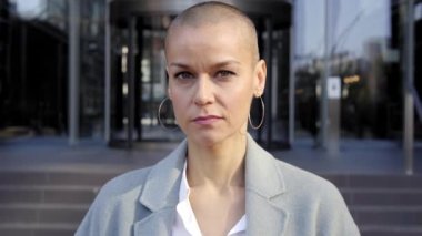 Tıraşlı kafalı, kameraya bakan ciddi, orta yaşlı yetkin bir kadın. Dışarıdaki iş adamları bina şirketleriyle birlikte..