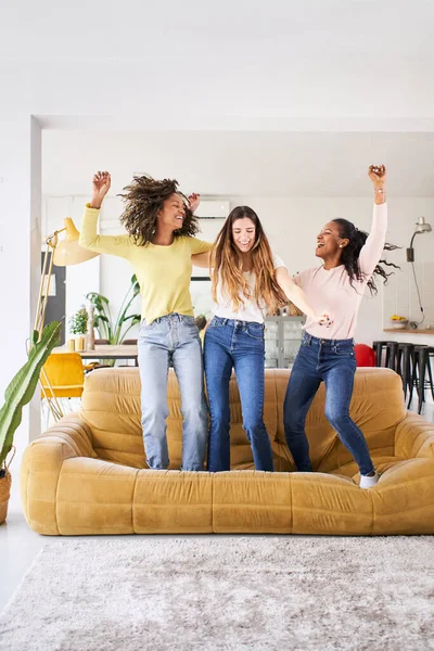 Вертикальне фото трьох смішних друзів-жінок, які танцюють на дивані в приміщенні. Багаторасові щасливі жінки грають і святкують радість життя Веселі дівчата насолоджуються разом вдома розважаються . — стокове фото