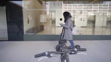 E- scooterlı akıllı telefon şehir caddesini kullanarak yürüyen bir iş kadını. Gençler, modern mimari geçmişe sahip sağlıklı bir yaşam tarzı ile eko taşımacılığı yapıyorlar..