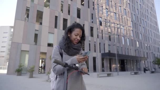 Feliz sorridente mulher de negócios andando usando telefone inteligente centro financeiro rua urbana da cidade com e-scooter. Jovens montam eco transporte saudável estilo de vida com arquitetura moderna fundo. — Vídeo de Stock