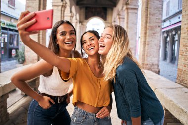 Üç neşeli kız yaz kıyafetleri içinde turistik şehir merkezinde selfie çekiyorlar.