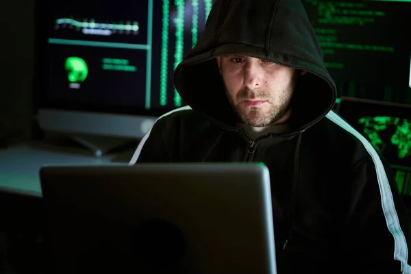 Portrait of criminal hooded hacker sitting at desk and breaking into government or big company data servers in dark atmosphere with system codes animation in background. — Foto de Stock