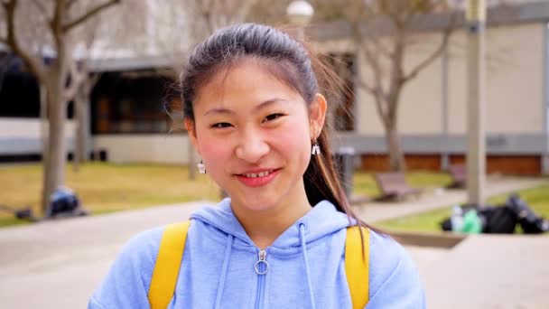 Chinese teenager smiling happy at university campus. Close up portrait of cheerful Asian girl looking at camera standing outdoors on blurred background. — Vídeo de stock